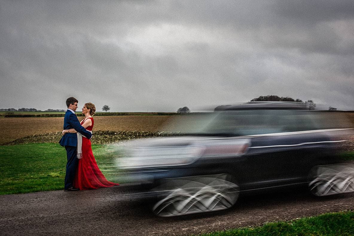 bride and groom hugging as car drives by Kingscote Barn
